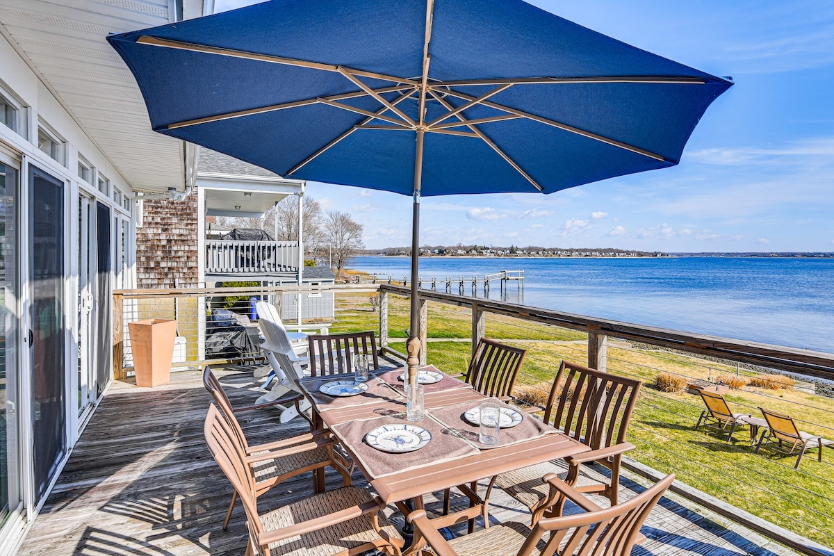 An outdoor dining area features a large table surrounded by wooden chairs, set under a blue umbrella. The space offers views of the bay, with a grassy area and lounge chairs visible in the foreground. The shoreline is seen in the distance.