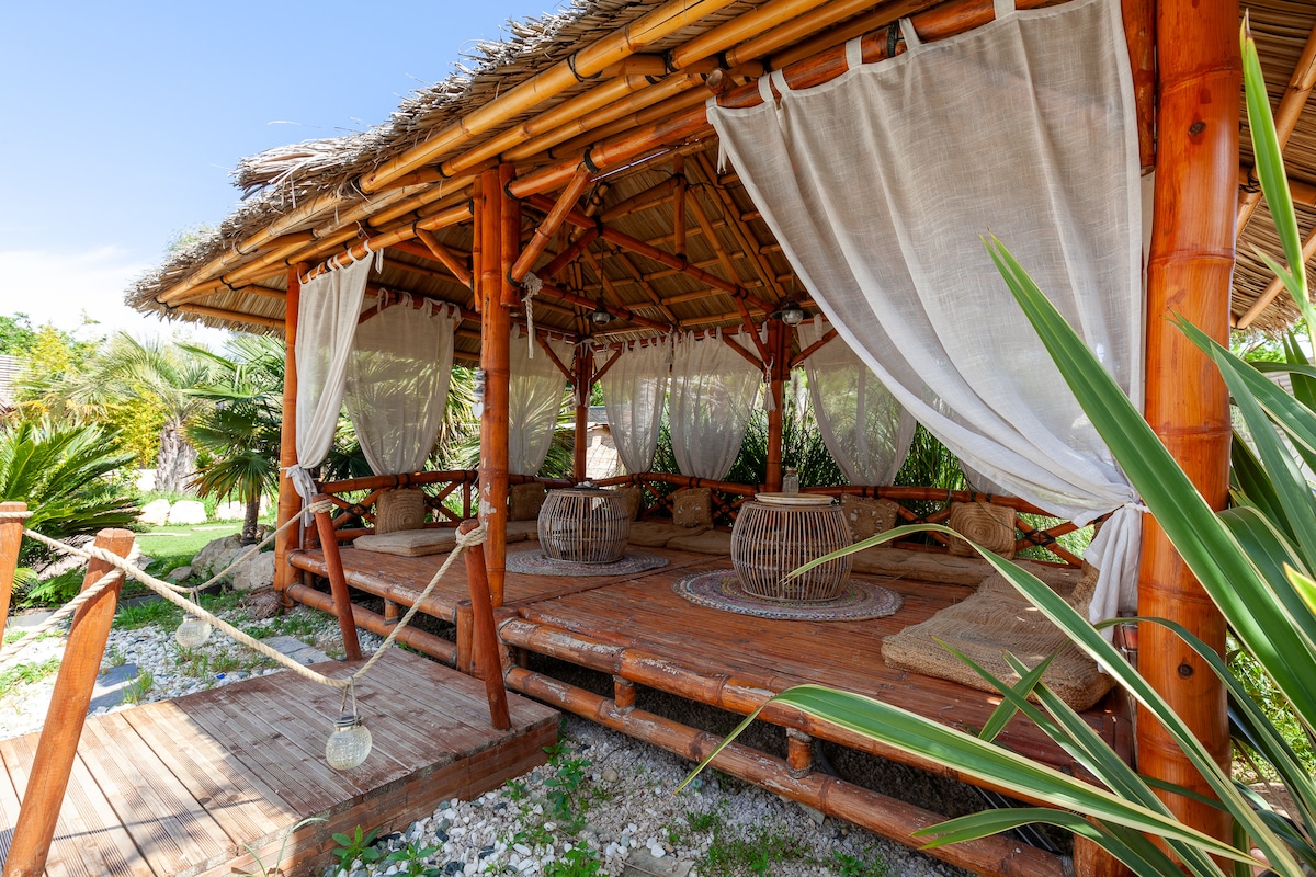 A shaded outdoor lounge area features a wooden structure with bamboo accents and flowing curtains. Comfortable seating is arranged on a wooden deck adorned with natural textures, surrounded by lush greenery and plants, creating an inviting space for relaxation.