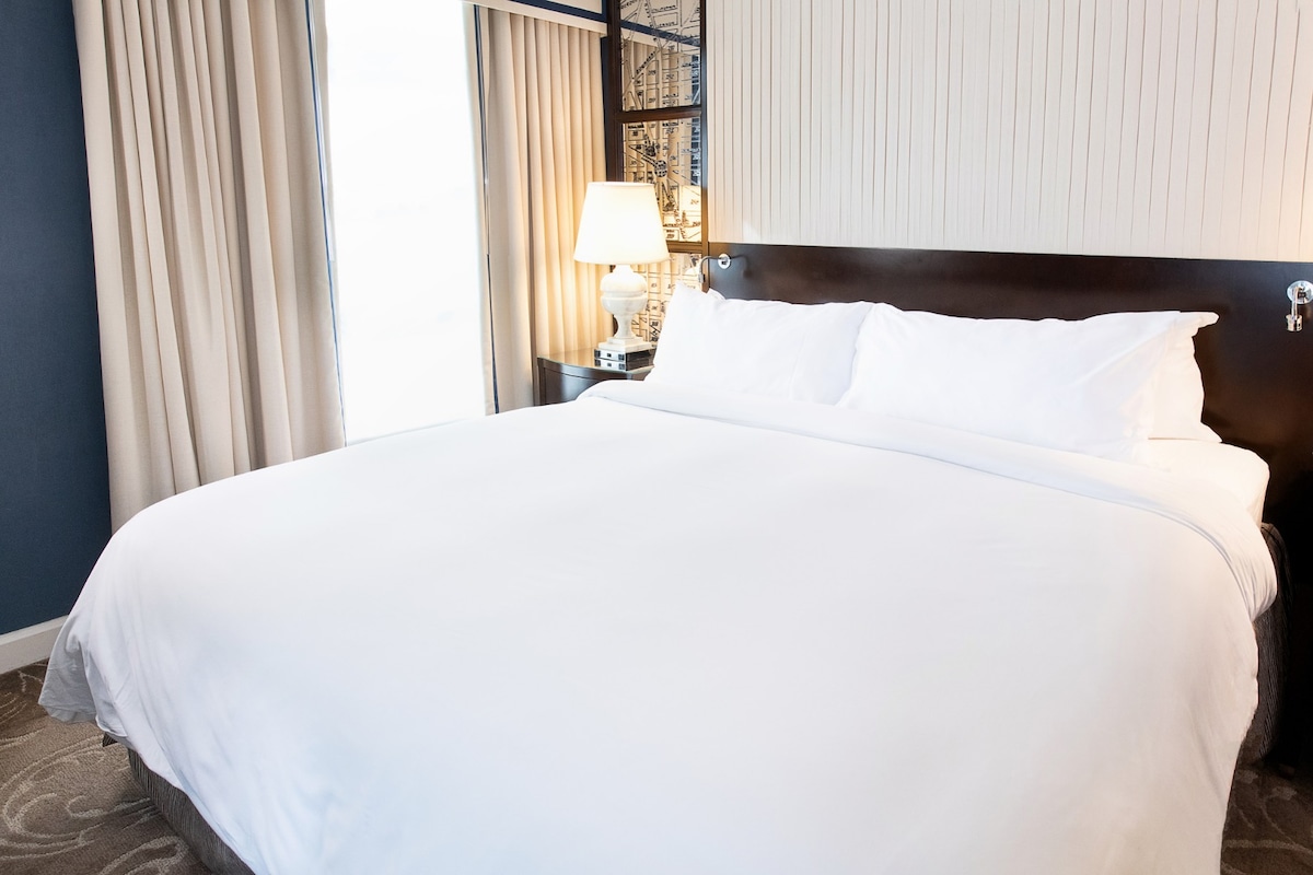 A spacious king bed is centered against a backdrop of soft, neutral tones, adorned with crisp white linen. A sleek bedside lamp provides gentle illumination, while large windows dressed with light curtains allow for natural light to fill the room.
