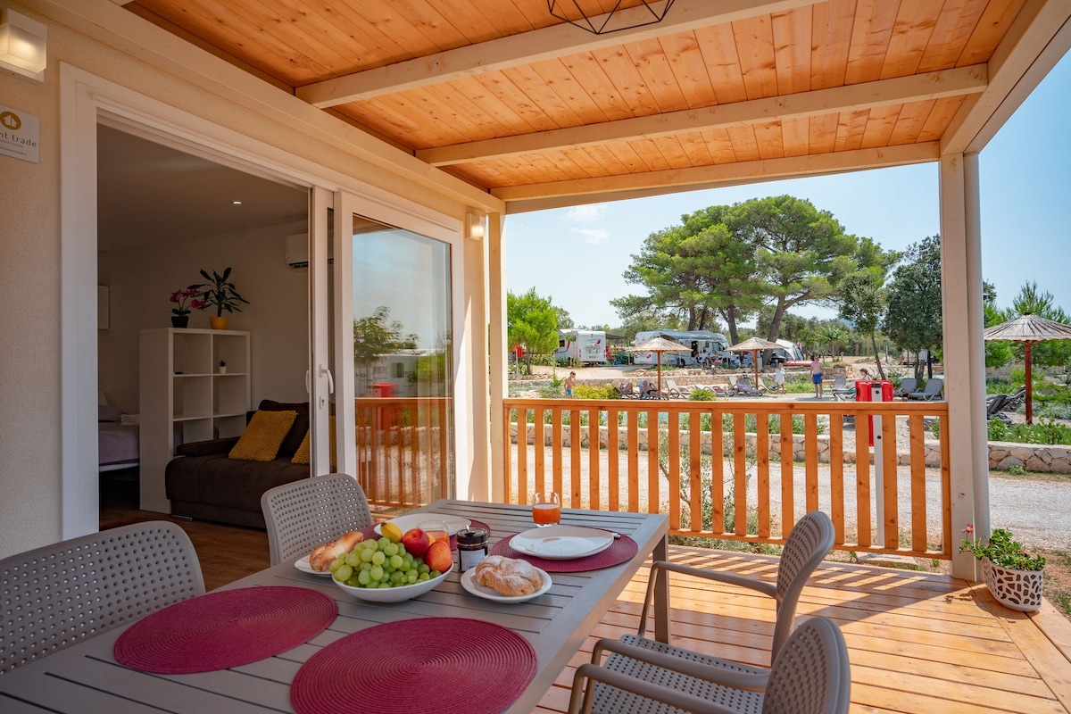 An outdoor terrace is shown, featuring a dining table set with fruits and pastries. The space is shaded by a wooden roof, overlooking a landscaped resort area with greenery and sun umbrellas in the distance. Natural light enhances the inviting atmosphere of the terrace.