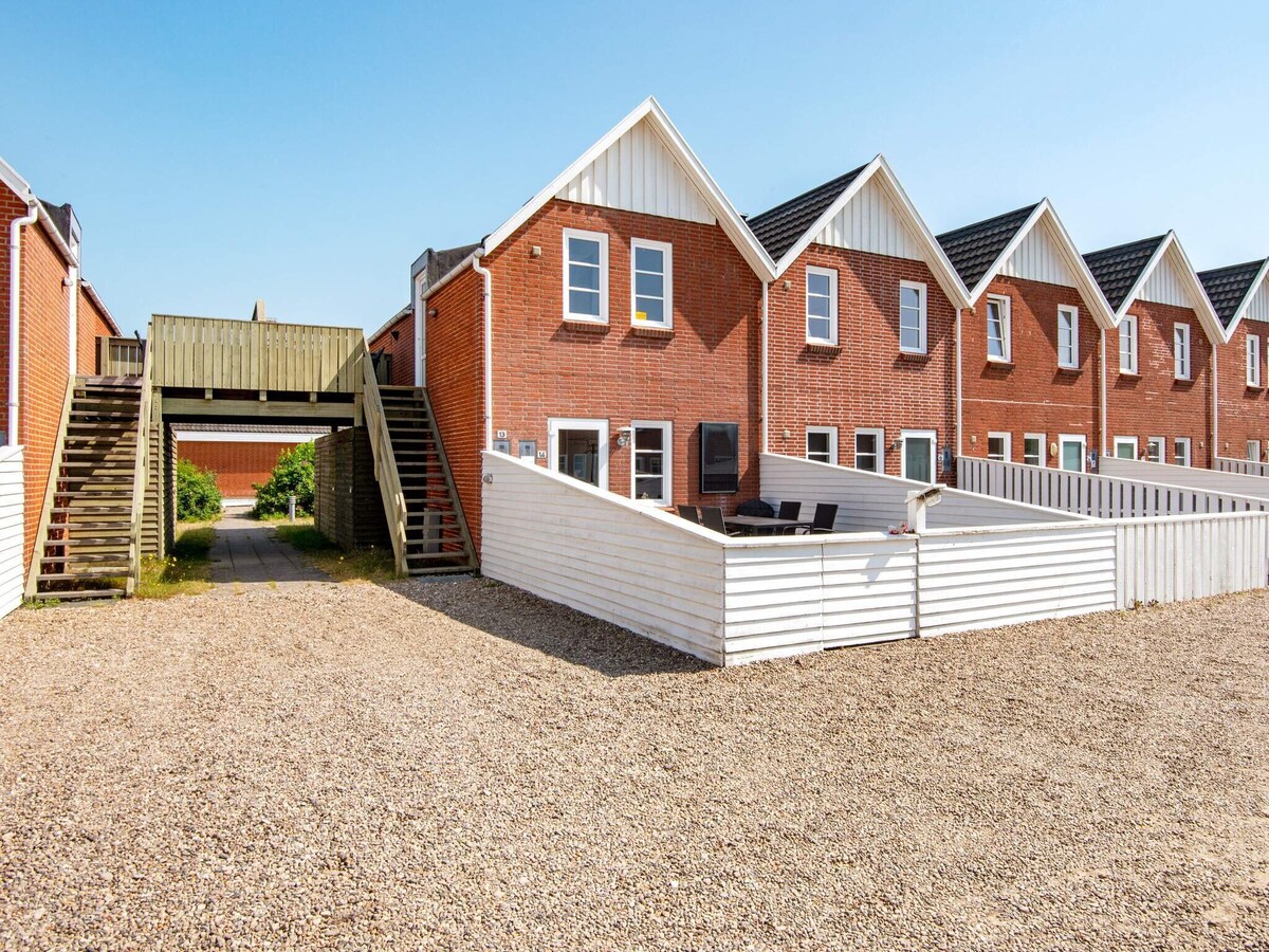A row of holiday homes is presented, featuring red brick exteriors and gabled roofs. A sheltered terrace with outdoor furniture is accessible from the first floor. Pathways and staircases provide connectivity between units, and a gravel area surrounds the premises.