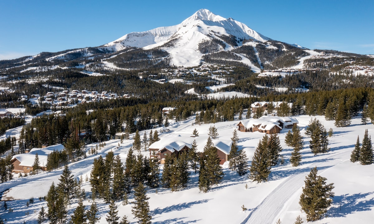 The image captures a winter landscape with a snow-covered valley and a backdrop of majestic mountains. Charming homes can be seen nestled among snow-dusted trees, while ski trails are visible on the slopes in the distance, providing a picturesque view of the serene setting.