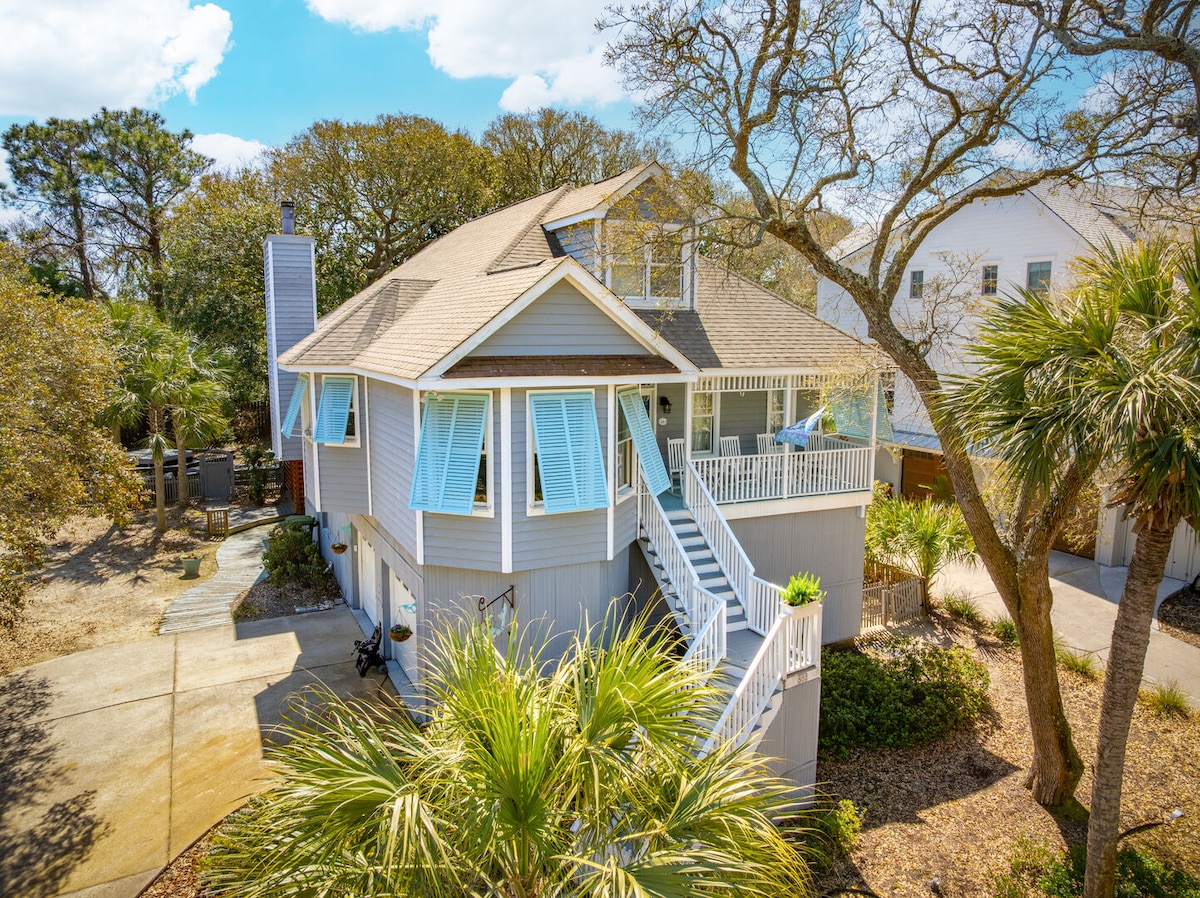 A spacious three-story beach home is portrayed, surrounded by lush trees and tropical landscaping. The exterior showcases light-colored siding and a large deck accessed by stairs, with ample outdoor seating visible. Windows are adorned with shutters, contributing to the coastal charm of the residence.