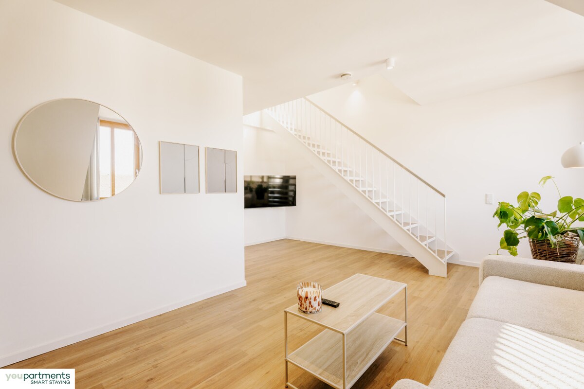 A spacious living area is highlighted by a large mirror and minimalistic wall art. A modern TV is mounted on the white wall. Natural light fills the room through large windows, complementing the wooden floor and the neutral-colored sofa, with a small coffee table placed in front.
