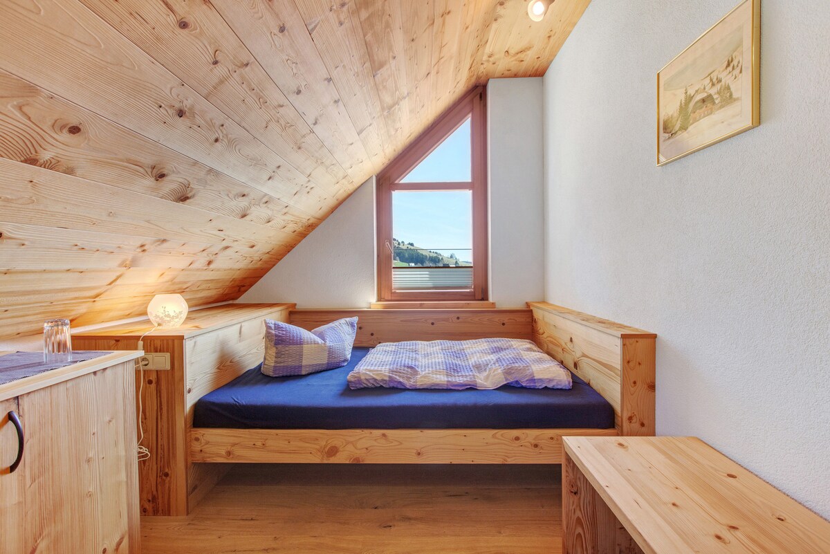A cozy single room features wood-paneled walls and a sloped ceiling, creating a warm atmosphere. There is a spacious bed with blue bedding, placed against the wall, and a compact table nearby. A window allows natural light to fill the room, revealing views of the surrounding mountains.