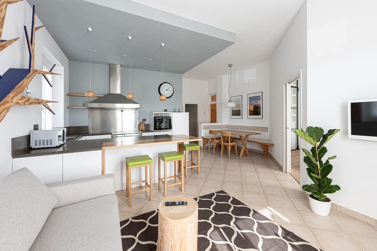 An open kitchen and living area combines functionality and comfort. The space features a large kitchen island with modern appliances, wooden stools, and a dining table. A plush sofa and a decorative plant enhance the inviting ambiance of the room.