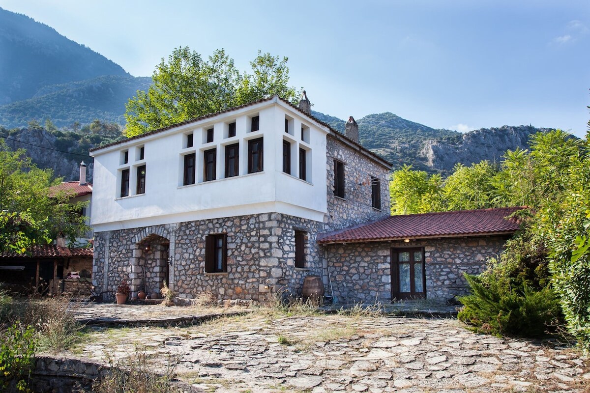A traditional stone house is surrounded by greenery and mountainous terrain. The structure features large windows that allow natural light to enter. The exterior showcases a blend of stone and white finishes, complemented by a terracotta roof and a spacious outdoor area with natural landscaping.