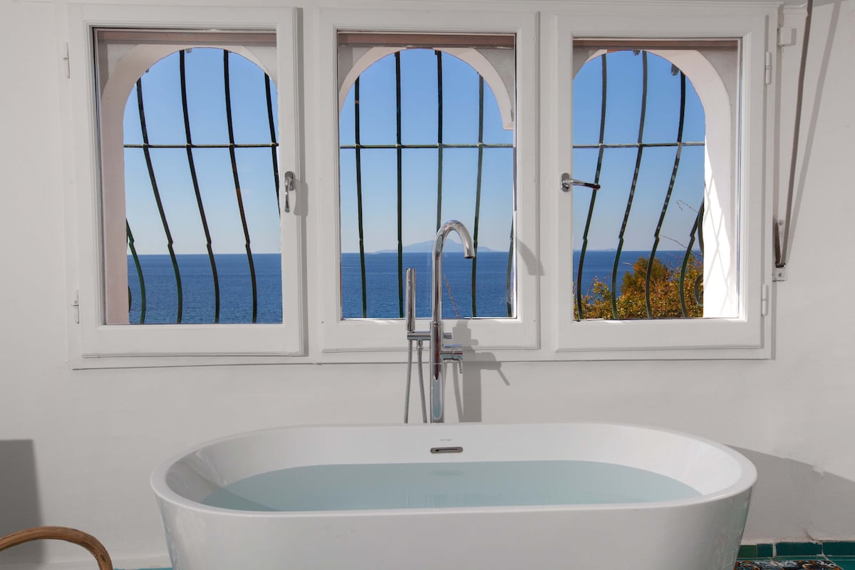 A freestanding bathtub is positioned in front of three arch-shaped windows, allowing for a panoramic view of the sea. The tranquil water reflects the natural light entering through the windows, with decorative grilles adding a unique architectural element.