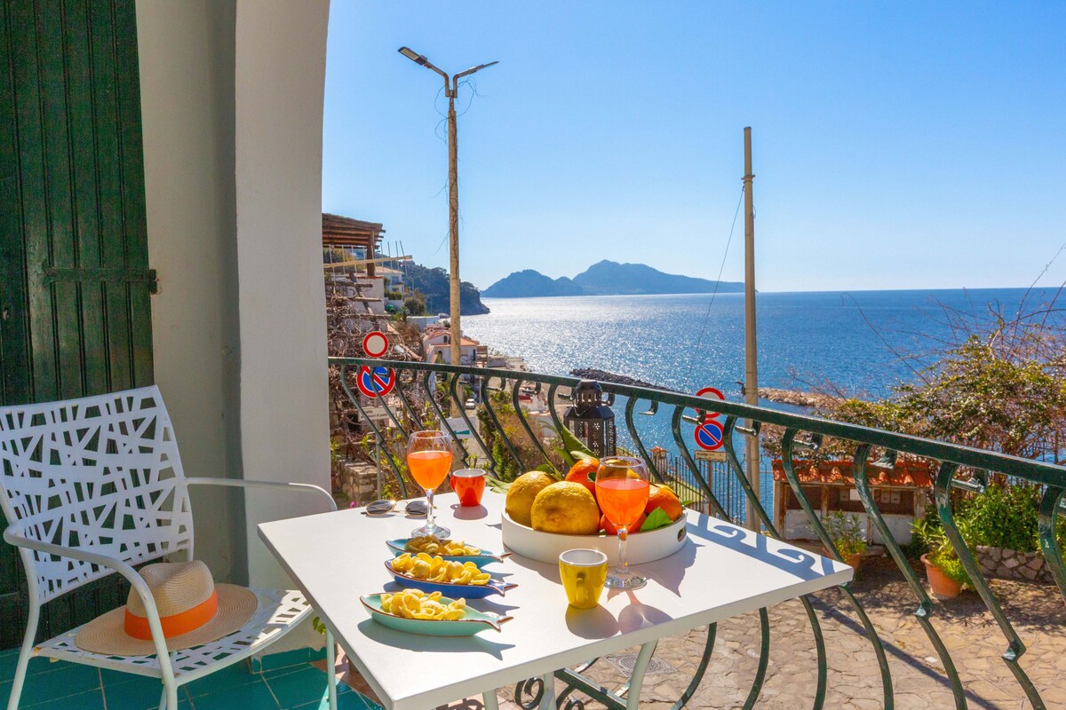 A small balcony features a table set with two colorful drinks and a plate of fruit, overlooking a serene sea view with distant mountains. Two light chairs frame the table, and sunlight brightens the scene against a clear blue sky.