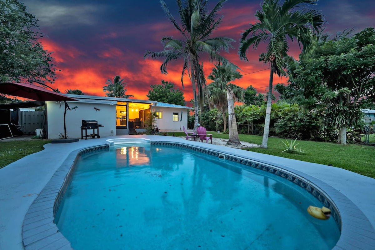 A serene private pool is framed by lush greenery, with palm trees gently swaying in the background. The vibrant sunset sky casts warm colors over the scene, creating a tranquil outdoor space perfect for relaxation.