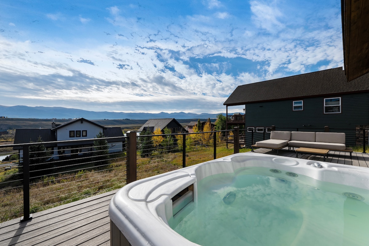 A private hot tub is situated on a spacious deck, surrounded by a wooden railing. Scenic views of the surrounding landscape and distant mountains unfold in the background, with hints of autumn foliage visible. Comfortable seating options are arranged nearby for relaxing outdoors.