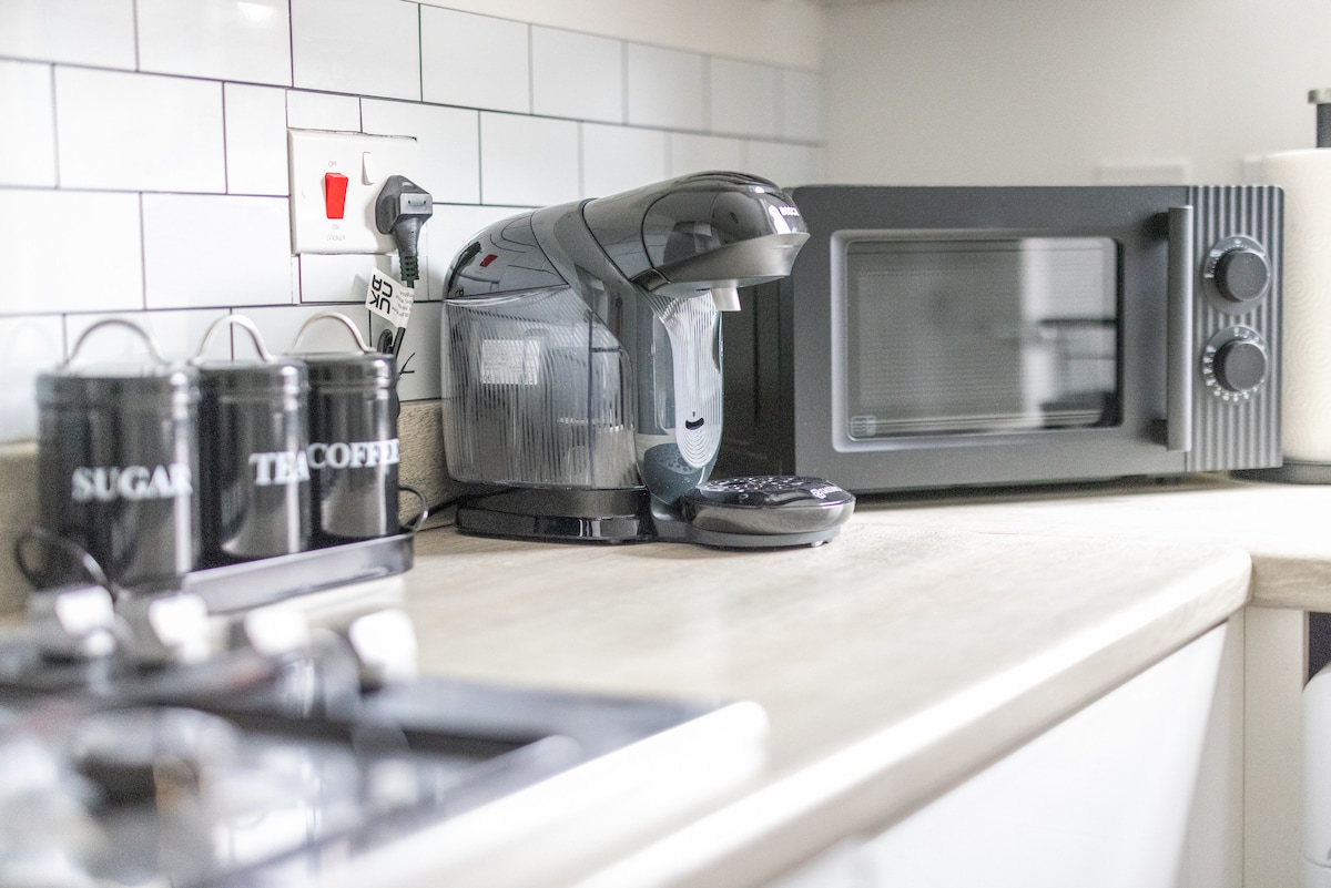 A modern kitchen setup is displayed, featuring a coffee machine, a microwave, and containers labeled for sugar, tea, and coffee. The light countertop provides a bright contrast against the dark appliances, and the clean tiled backsplash adds a contemporary touch.