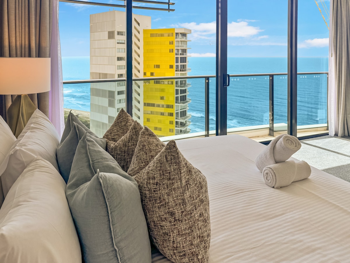 A spacious bedroom features a large bed adorned with several decorative pillows. Soft, neutral bedding complements the room's calming aesthetic. A glass door opens to a balcony, revealing expansive views of the ocean and the skyline, enhancing the inviting atmosphere.