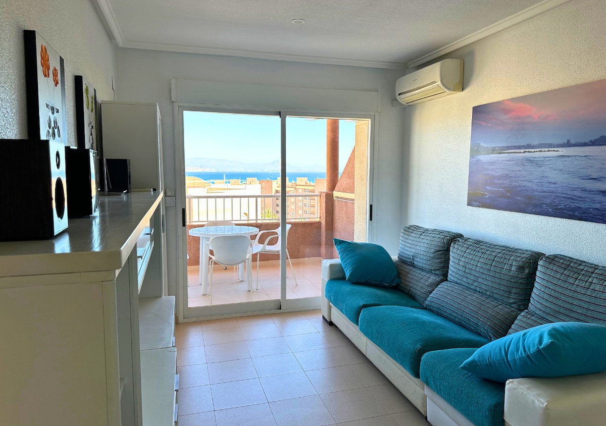 A bright living space includes a comfortable sofa with teal accents and a view of the ocean through large sliding glass doors. A small dining table and chairs can be seen on the private terrace, providing an inviting space for relaxation.