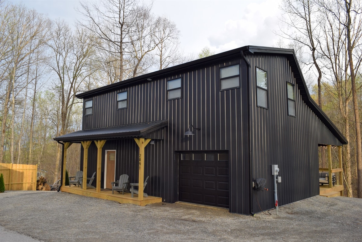 A modern two-story house features a sleek black exterior, complemented by wooden accents. The front porch includes several outdoor chairs, providing a welcoming entrance. Large windows enhance the building's design, while the surrounding trees create a natural backdrop, emphasizing the peaceful setting.