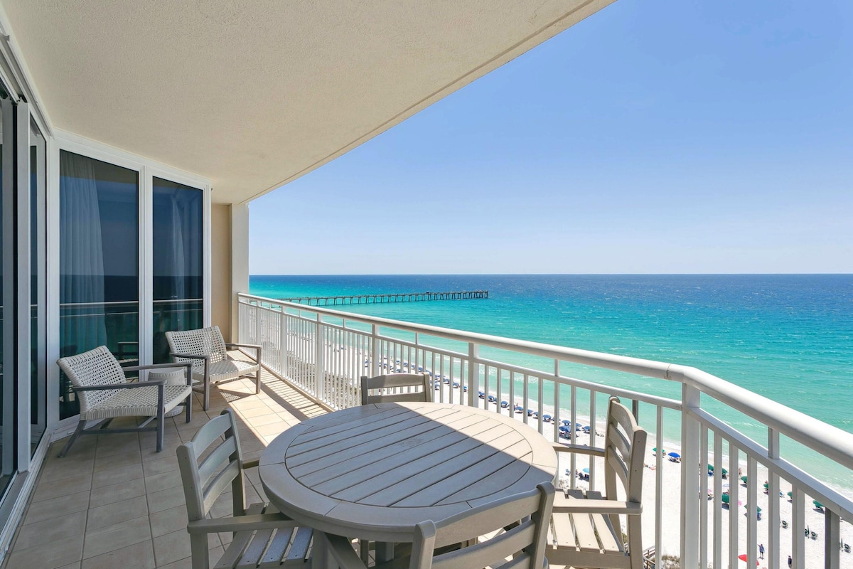 An expansive balcony with a round table surrounded by comfortable chairs offers stunning views of the Gulf of Mexico. The turquoise waters gently meet the shore, while the distant pier adds a scenic touch to the beach landscape.