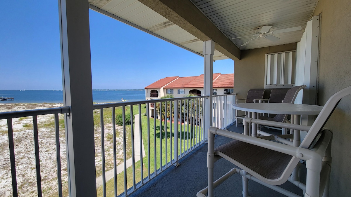 A private balcony is shown with a view of the Intracoastal Waterway. Comfortable outdoor seating includes a table with four chairs. The surrounding area features grassy grounds and nearby buildings, creating a serene setting for relaxation.