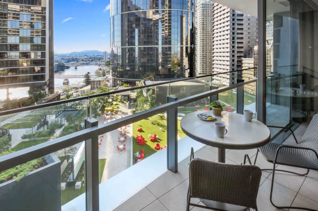 A private balcony features a small round table accompanied by two chairs, overlooking a well-maintained garden area below. Modern buildings and the Brisbane skyline serve as a backdrop, with sunlight illuminating the space and panoramic views of the surrounding cityscape.