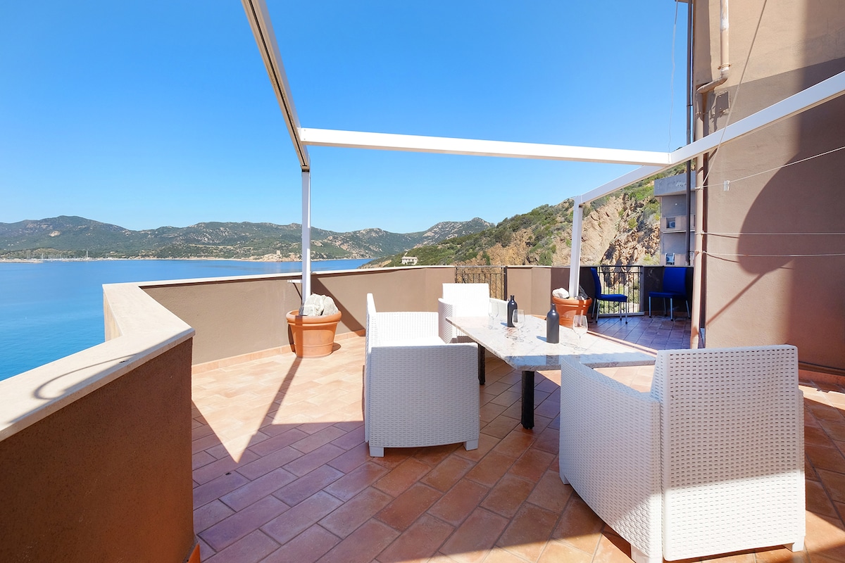 An expansive terrace is showcased, featuring a dining table surrounded by wicker chairs. Panoramic views of the sea and hills are visible in the background. Sunlight bathes the area, highlighting the terracotta tiles and the light-colored exterior wall.