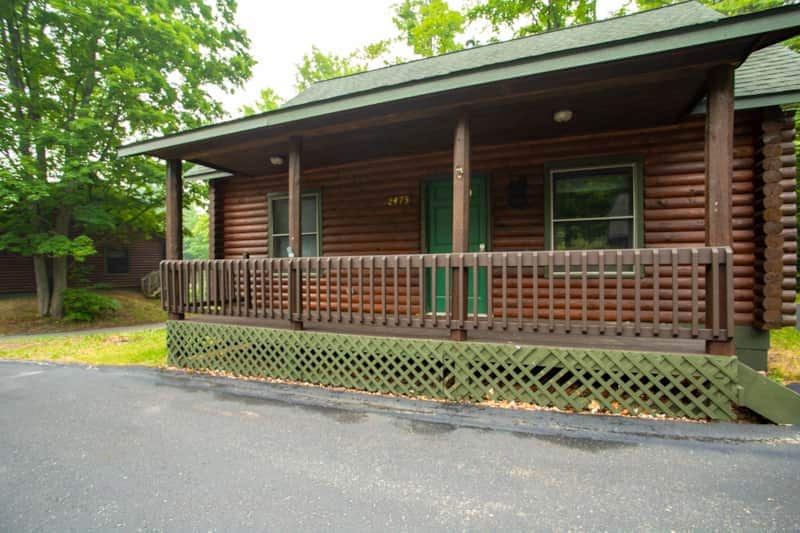 The exterior of the log cabin displays a welcoming porch with wooden railings and green doors. The entrance is framed by lush trees, providing natural shade. The cabin's rustic design features wooden siding and is situated on a paved pathway.