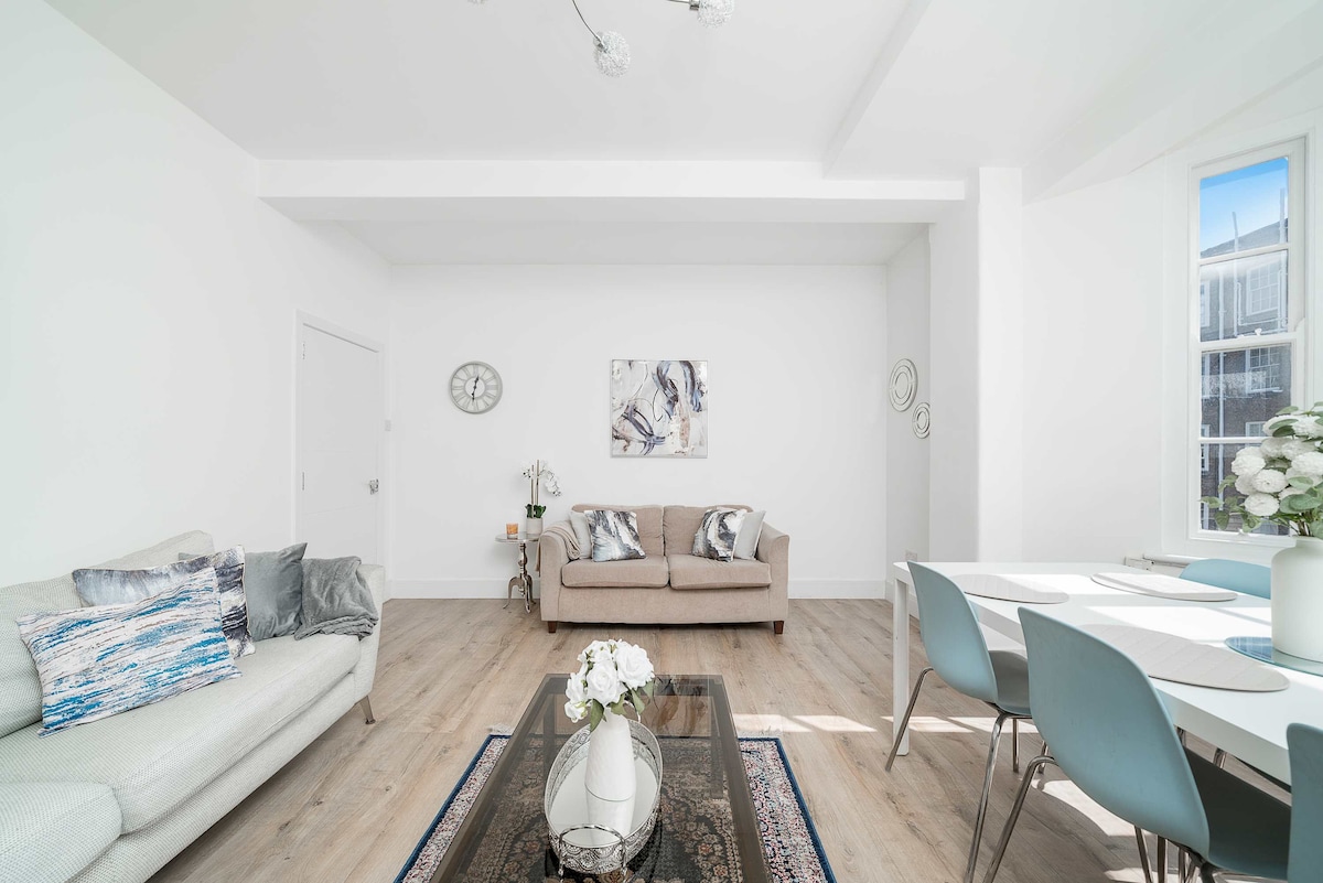 A bright living area is showcased, featuring a light-colored sofa and a second seating arrangement against a bright white wall. A decorative table occupies the center, surrounded by a dining table with blue chairs. Natural light enters through large windows, enhancing the open feel of the space.