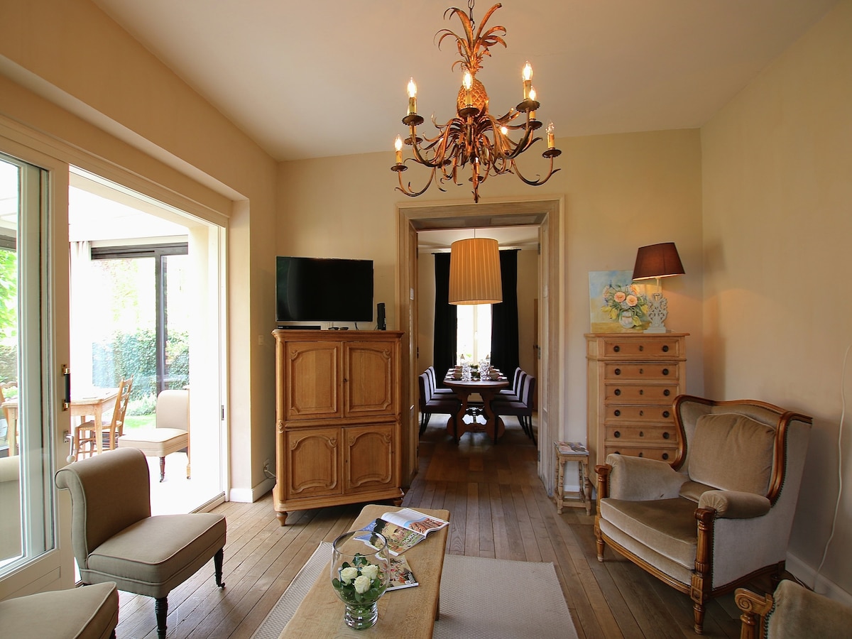 A welcoming living area features a combination of wooden flooring and neutral-toned walls. An ornate chandelier hangs above the seating arrangement, which includes an armchair and a sofa. A rustic cabinet and a flat-screen TV provide additional amenities, while large glass doors lead to a conservatory.