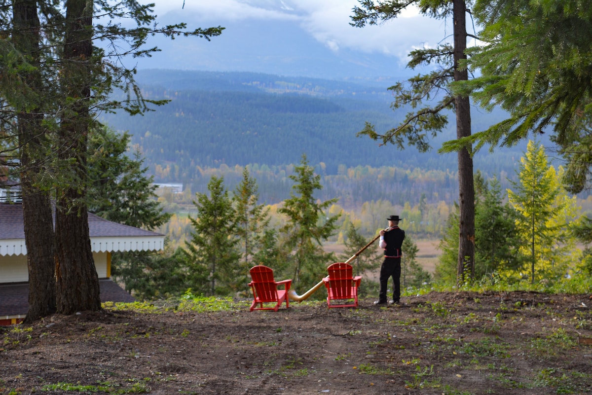 A scenic view from a hillside features two red Adirondack chairs positioned on a grassy area. In the background, rolling hills and dense forests extend towards the horizon under a partly cloudy sky. A figure dressed in a traditional outfit stands beside the chairs, holding a rope.