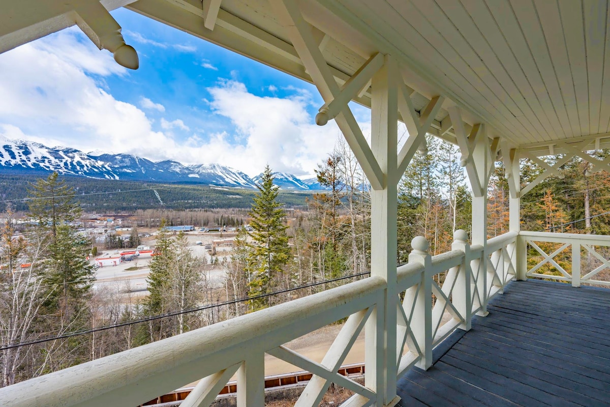 A spacious balcony offers striking views of the surrounding mountains and valley. Wooden railings and supports create an inviting space for relaxation. The natural scenery features a mix of evergreen trees and distant snow-capped peaks under a blue sky with scattered clouds.