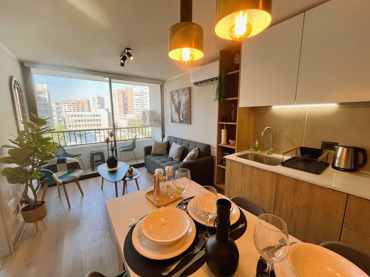 A modern living space features a dining table set for four, complemented by stylish tableware. The inviting lounge area includes a grey sofa with decorative pillows and a cozy throw. Natural light flows through large windows, highlighting the well-equipped kitchen area in the background.