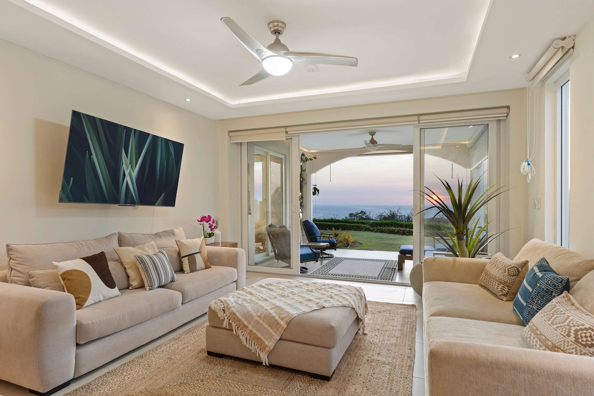 A modern living room is characterized by plush beige sofas with decorative pillows and a central ottoman. A large flat-screen TV is mounted on the wall, while floor-to-ceiling glass sliders lead to a furnished patio. Soft light illuminates the space, highlighting views of the ocean and sunset.
