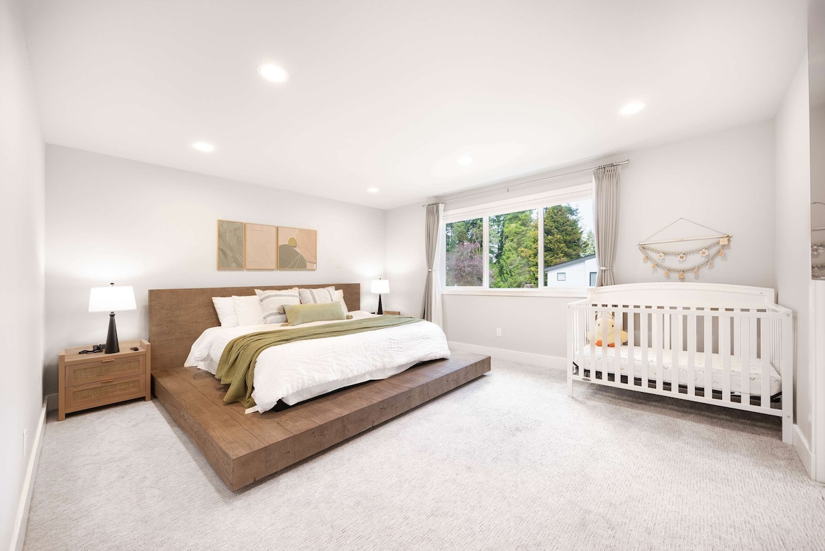 Una espaciosa cama tamaño king se encuentra en una plataforma elevada, con una cuna adyacente, bañada de luz natural para un refugio familiar sereno y acogedor. 