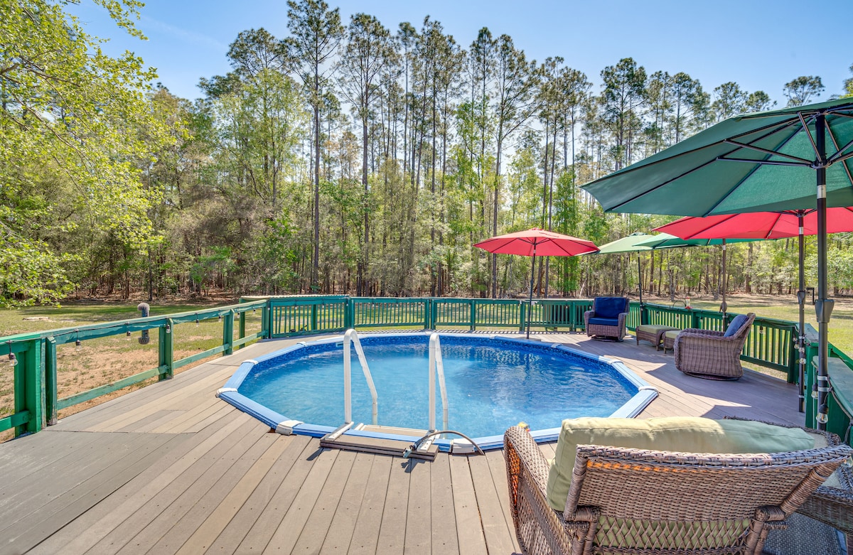 An outdoor pool is framed by a wooden deck, surrounded by lush trees. Colorful umbrellas provide shade over seating areas, including a wicker chair and a lounge. The serene natural setting offers a sense of privacy and tranquility.