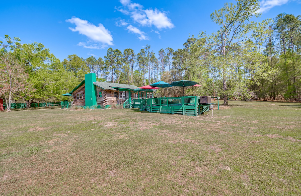 The cabin's exterior is surrounded by expansive green grass and forested areas. A spacious deck features colorful umbrellas and comfortable seating, creating an inviting outdoor space. The log cabin design is complemented by a vibrant green chimney, contributing to a natural and serene setting.