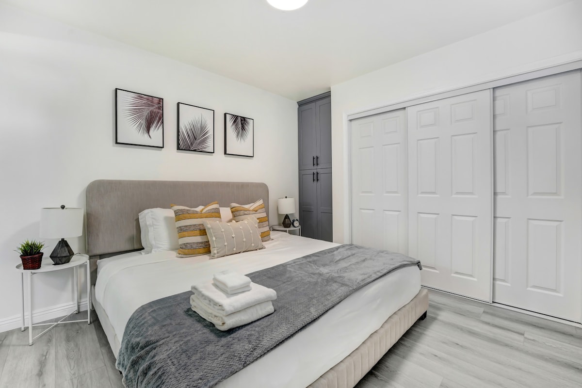 A light-filled bedroom features a spacious bed with layered bedding and decorative pillows. Art prints adorn the wall above the headboard, while a nightstand with a lamp is positioned beside the bed. A closet with sliding doors provides storage, completing the serene space.