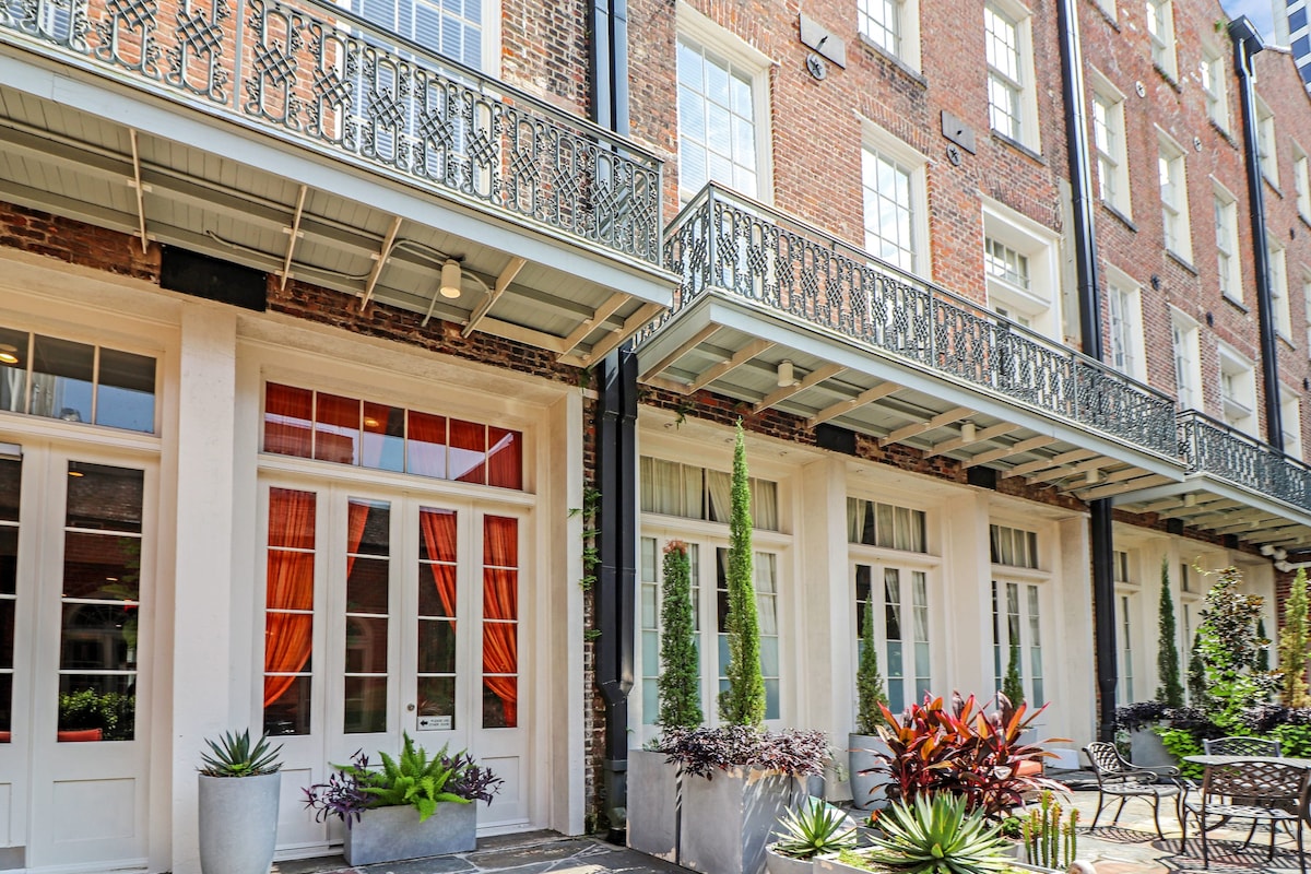 The exterior showcases a historic brick building with elegant wrought-iron balconies and large glass doors. Lush greenery and colorful plants enhance the inviting courtyard, while the vibrant curtains add a pop of color to the scene.