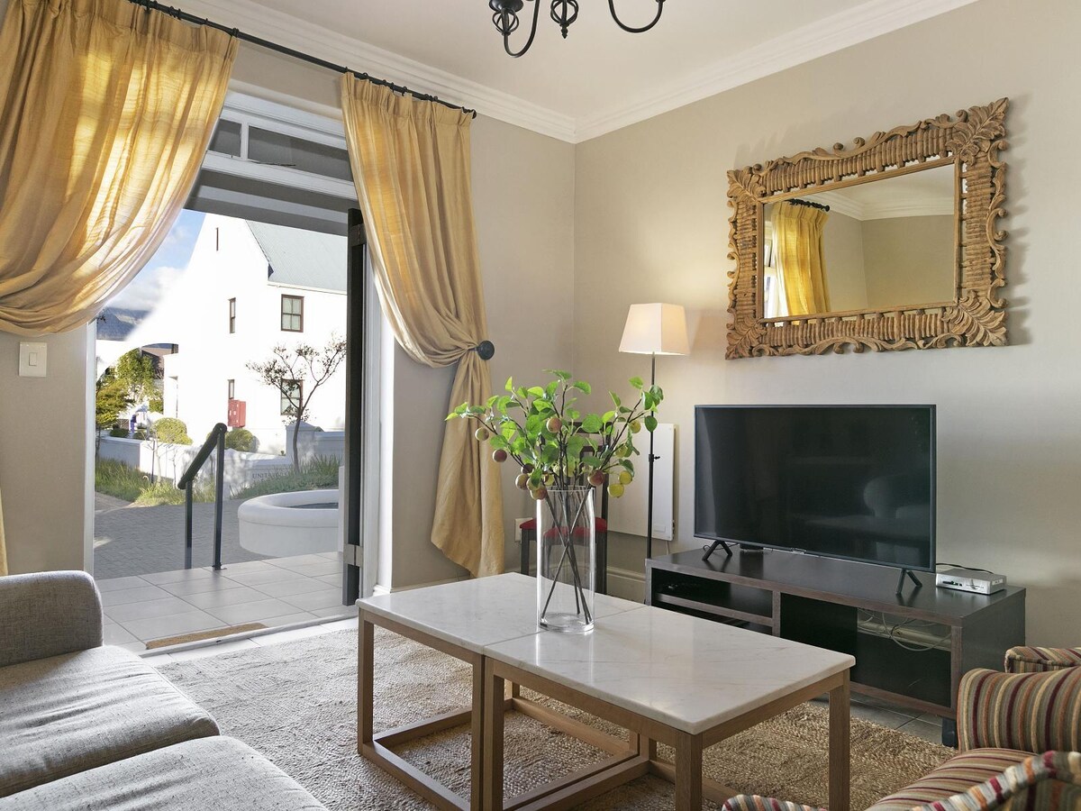 A comfortable living area features a large television placed on a low wooden console. A coffee table sits centrally, accompanied by a light-colored rug beneath. Elegant curtains frame the glass doors, allowing natural light to enter and offering views of the patio and outdoor space.