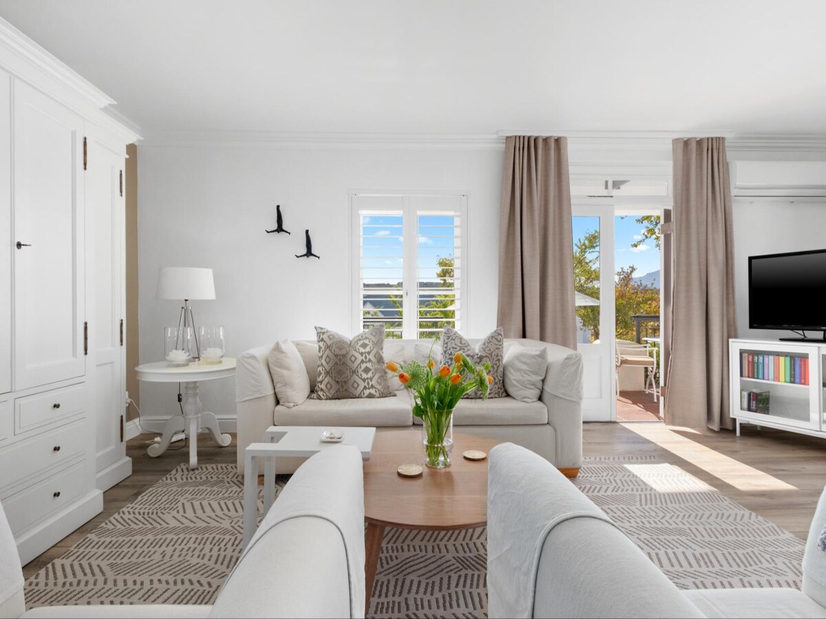 A bright living area features a light-colored sofa and armchairs arranged around a round coffee table with a floral centerpiece. Large windows provide natural light, while beige curtains frame the opening leading to a furnished patio. A flat-screen TV is positioned on a white media unit.