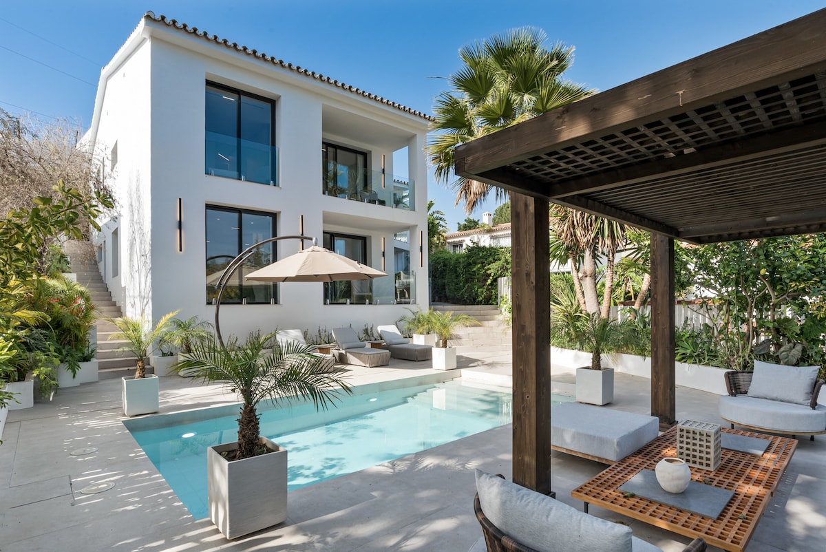 A modern villa features a heated pool surrounded by landscaped gardens and loungers. A shaded seating area with cushions offers a perfect spot for relaxation. The facade of the villa showcases large windows and a blend of white and natural materials.