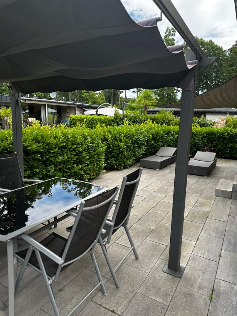 An outdoor dining area features a glass-top table surrounded by four metal chairs under a shaded canopy. Nearby, two loungers are positioned on a stone patio, bordered by lush green shrubbery. The tranquil environment invites relaxation and outdoor dining.