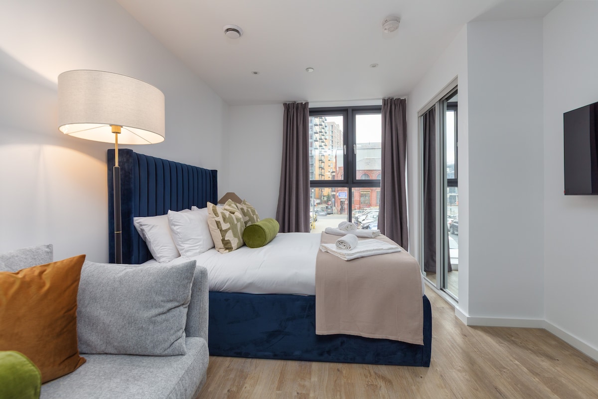 A well-lit bedroom features a spacious bed with decorative cushions and a neutral throw. A comfortable gray sofa is positioned beside the bed. Natural light enters through large windows with gray curtains, creating a cozy atmosphere. A television is mounted on the wall opposite the bed.