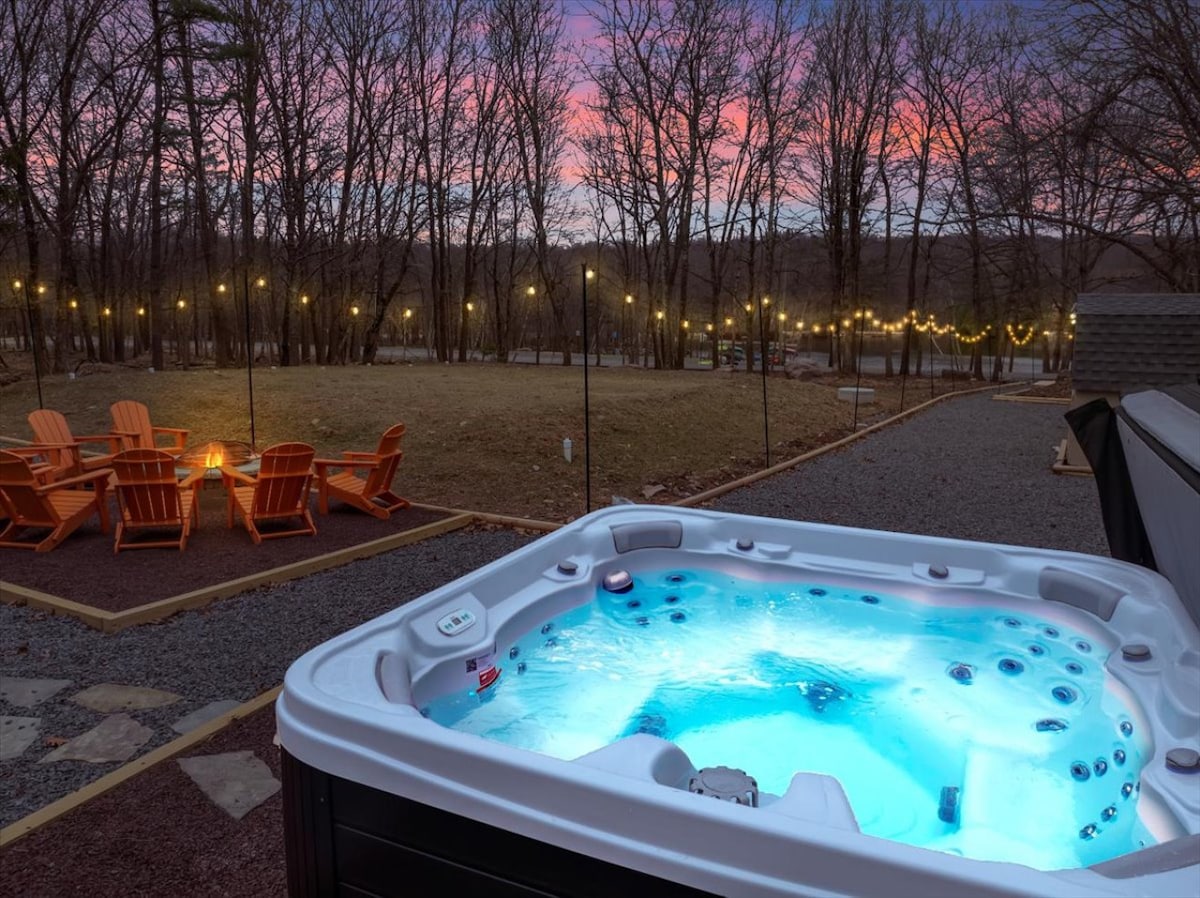 A private hot tub is surrounded by soft lights and a tranquil natural setting. The yard features Adirondack chairs positioned around a fire pit, inviting relaxation under the evening sky. The vibrant hues of dusk are reflected in the water of the hot tub.