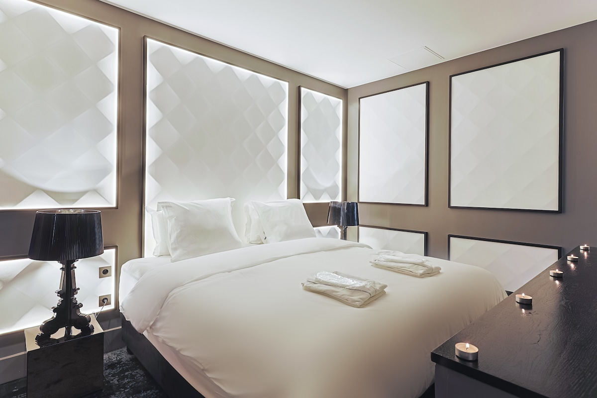 A serene sleeping area is highlighted by soft ambient lighting and textured wall panels. A spacious bed is adorned with neatly arranged towels, flanked by two black bedside lamps. The minimalist decor is complemented by simple framed artwork, creating a calm atmosphere.