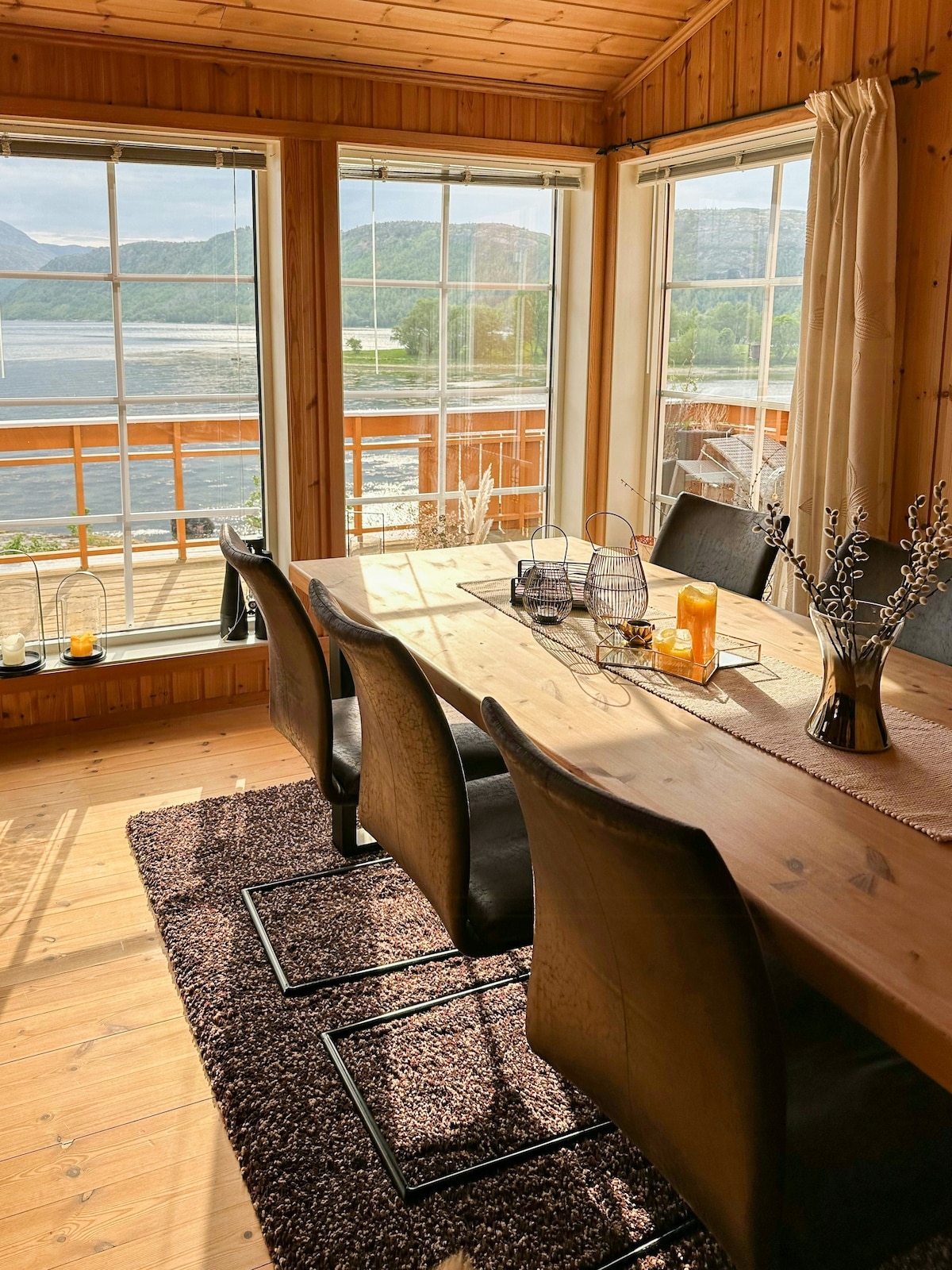A dining area features a large wooden table surrounded by six dark chairs, with natural light streaming in through large windows. Scenic views of the fjord and mountains are visible, enhancing the space's ambiance. A plush rug adds warmth to the wooden flooring.