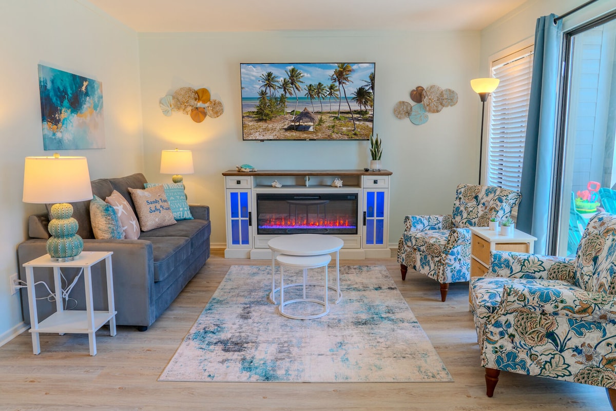 A welcoming living area features a comfortable gray sofa with decorative pillows, two patterned accent chairs, and a round coffee table. A wall-mounted flat-screen TV is above an electric fireplace, and natural light enters through sliding doors, enhancing the space's cozy ambiance.