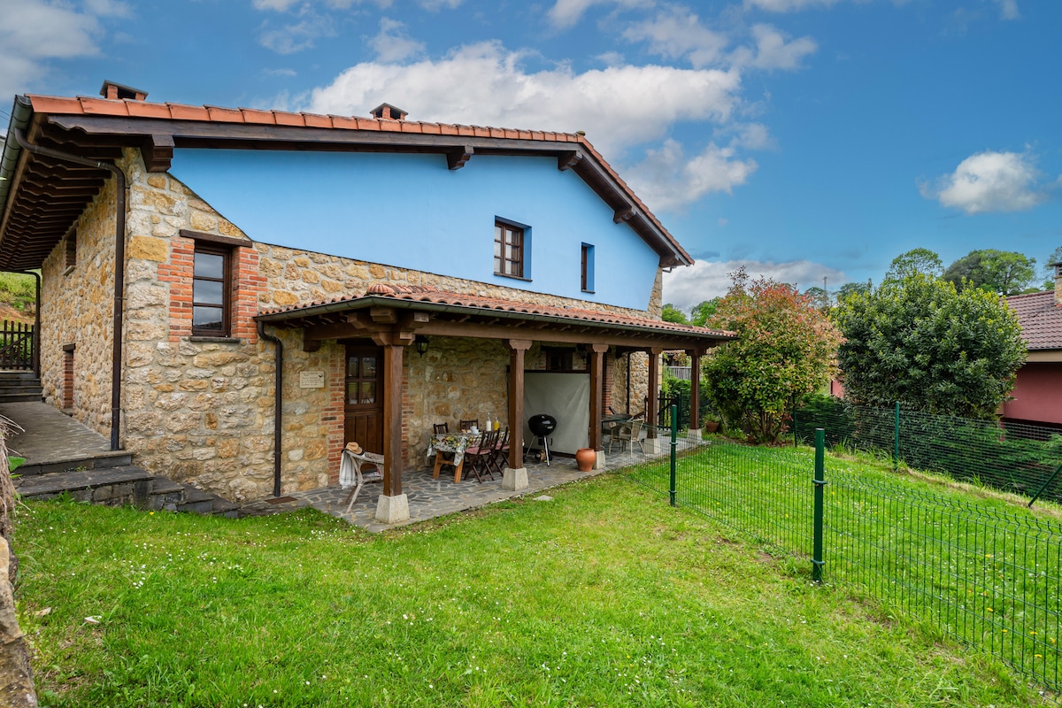 The exterior of the property showcases a charming stone facade with blue accents. A spacious private garden is surrounded by a fence, featuring a patio area with seating and a barbecue. Lush greenery blankets the ground, and trees can be seen in the background.