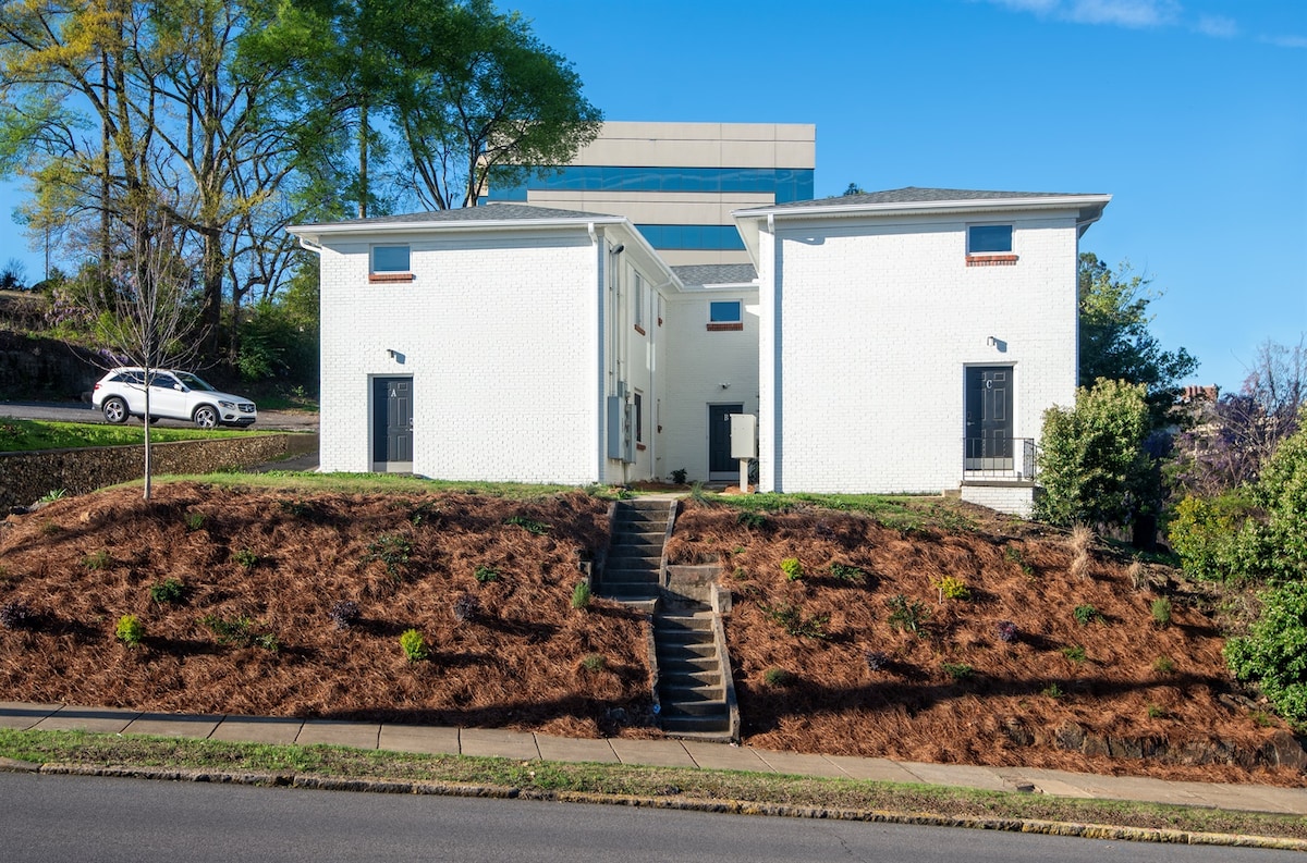 The exterior of a two-story building is shown, featuring a landscaped area with freshly laid mulch and small shrubs. A central stairway leads to the entrance, framed by individual entry doors for the units. A modern structure is visible in the background.