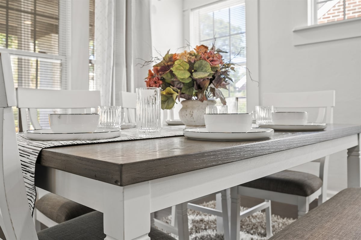 A light-filled dining area features a rustic wooden table set with simple white dishware. A floral arrangement adds a touch of color, complemented by glassware. Sunlight streams through large windows, illuminating the neutral-toned decor and enhancing the room's inviting atmosphere.