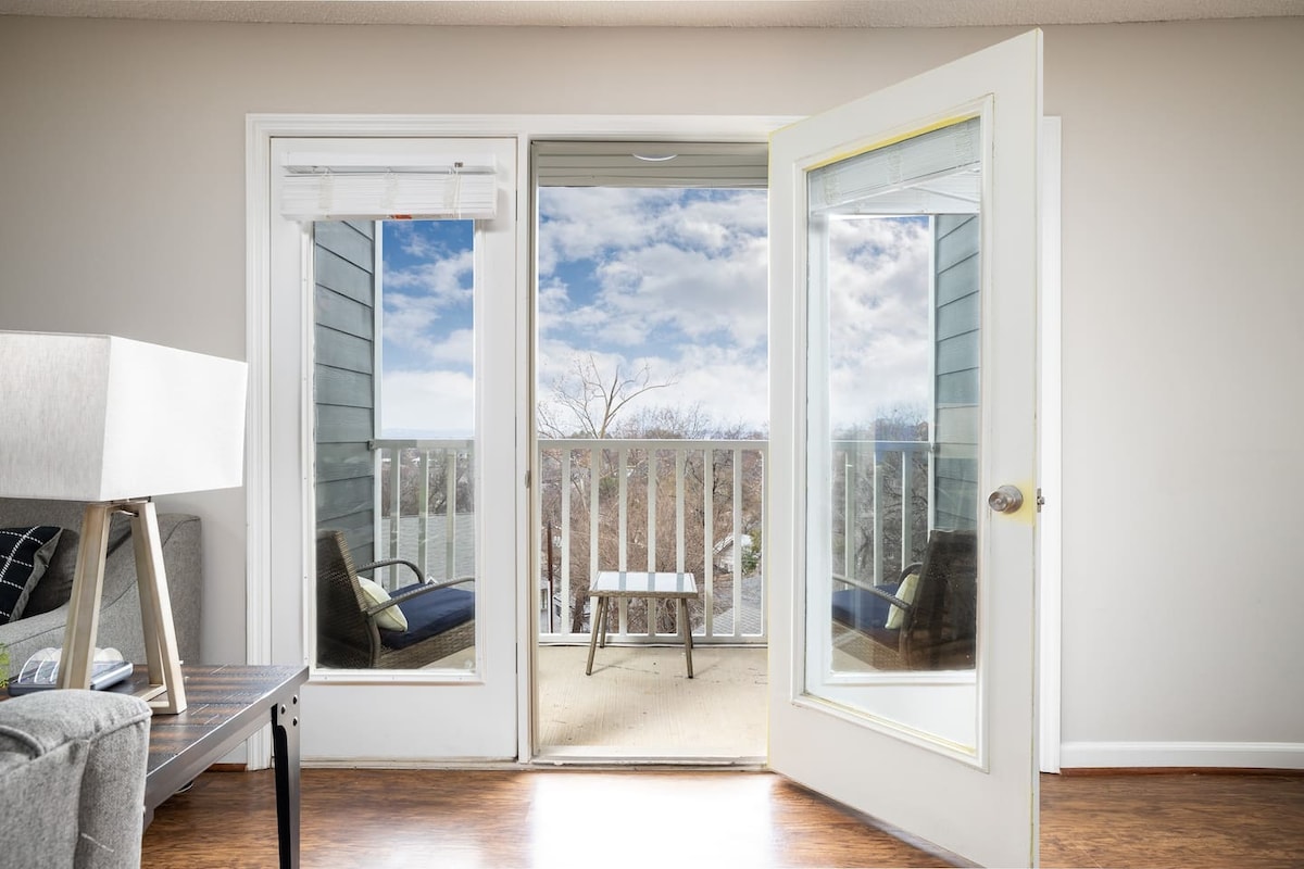 Double glass doors open to reveal a private balcony with outdoor seating, offering views of the surrounding landscape. Natural light floods the interior space, highlighting a cozy living area with a modern lamp and a coffee table.