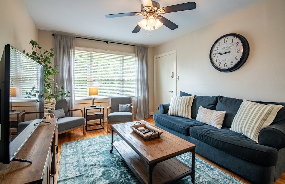A comfortable living room features a large dark sofa with decorative pillows and a wooden coffee table at its center. Natural light enters through large windows dressed with light curtains. Two armchairs and a floor lamp add additional seating, while a wall clock complements the decor.