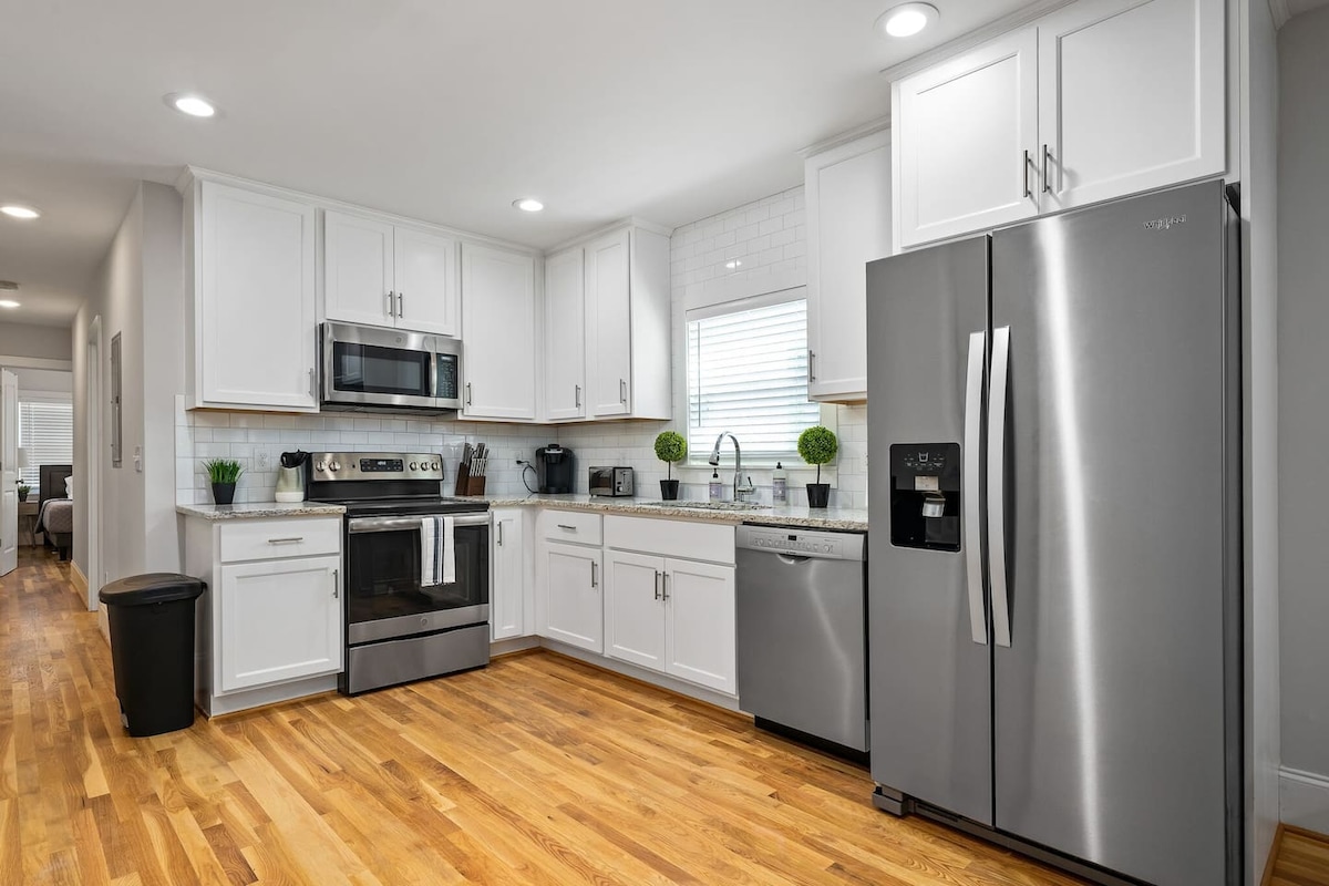 The kitchen features modern stainless steel appliances, including a refrigerator, microwave, and oven. White cabinetry contrasts with the hardwood floor, creating a bright and inviting space. Countertops are equipped with essential cookware and utensils, while small decorative plants add a touch of greenery.